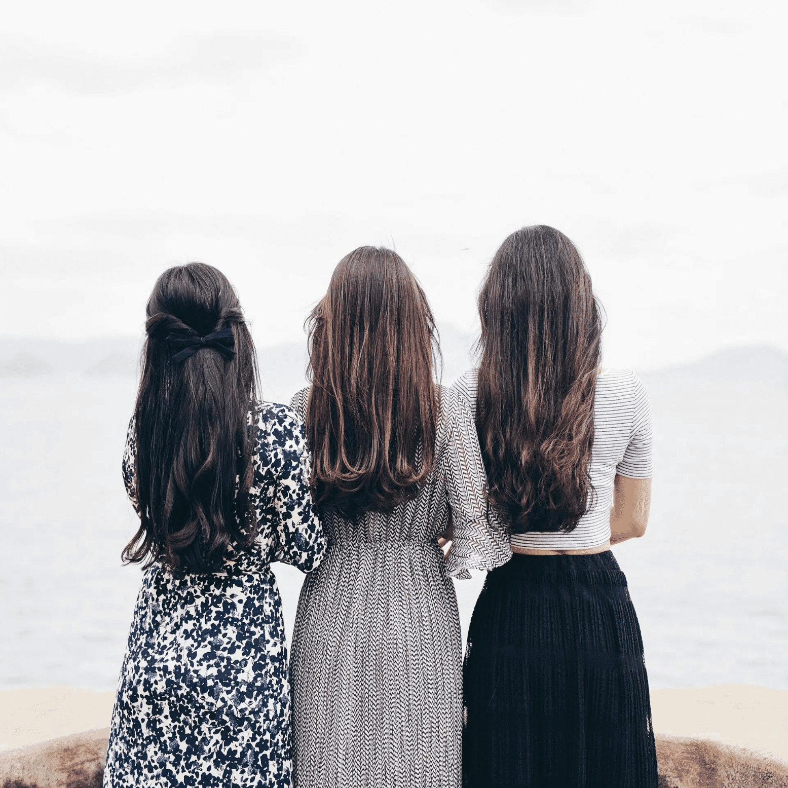 Three women with long hair standing by the sea, facing away.