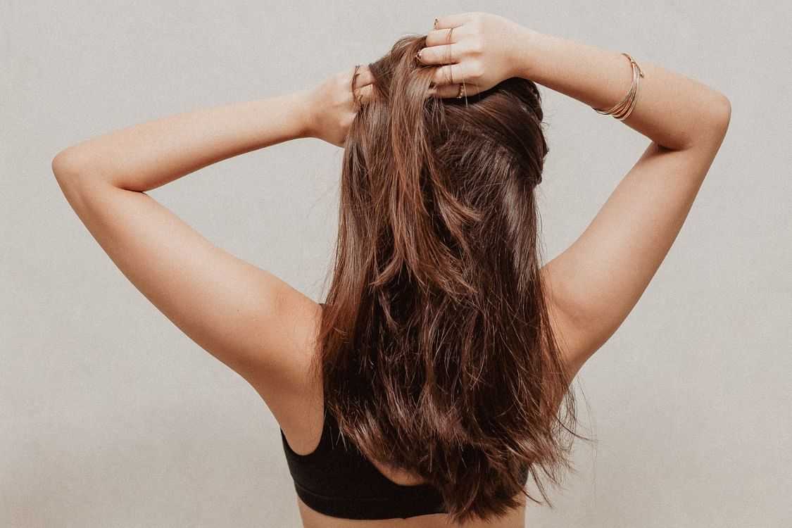 Woman tying her long hair up, shot from the back, neutral backdrop.
