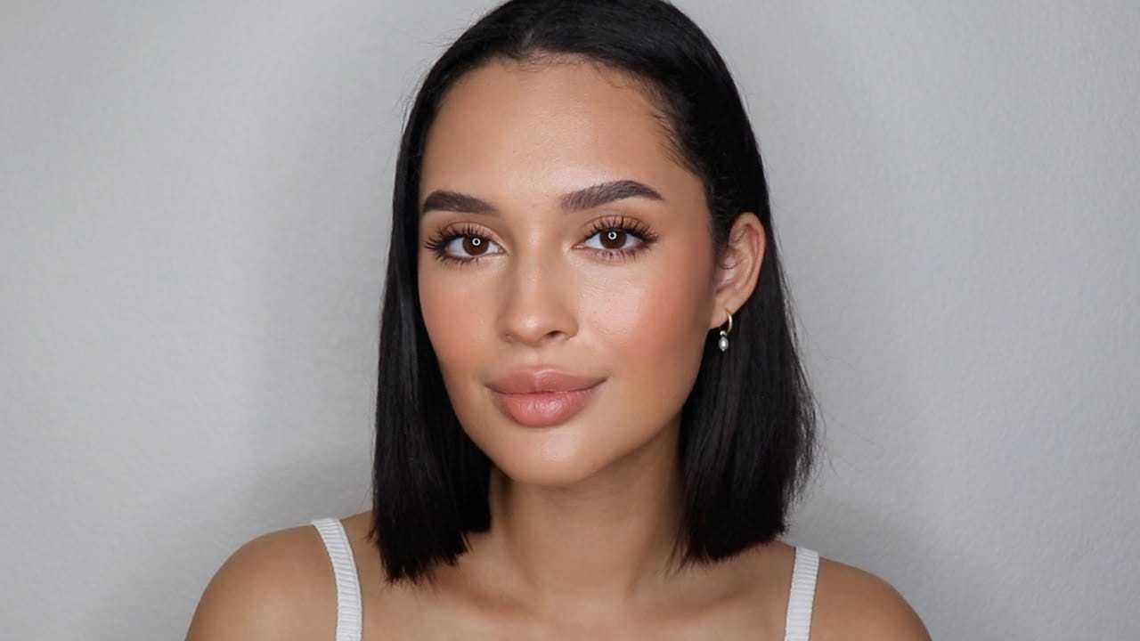 Woman with straight black hair and neutral makeup, wearing a white top and small earrings.